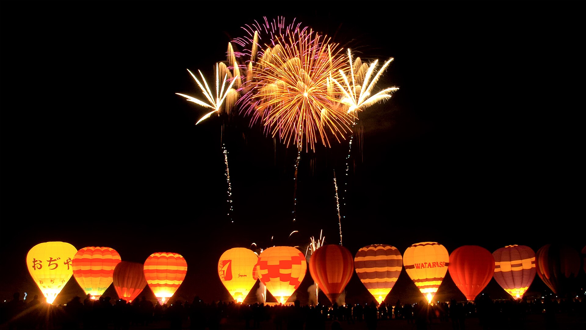 Hot air balloon night glow with fireworks over a festival crowd